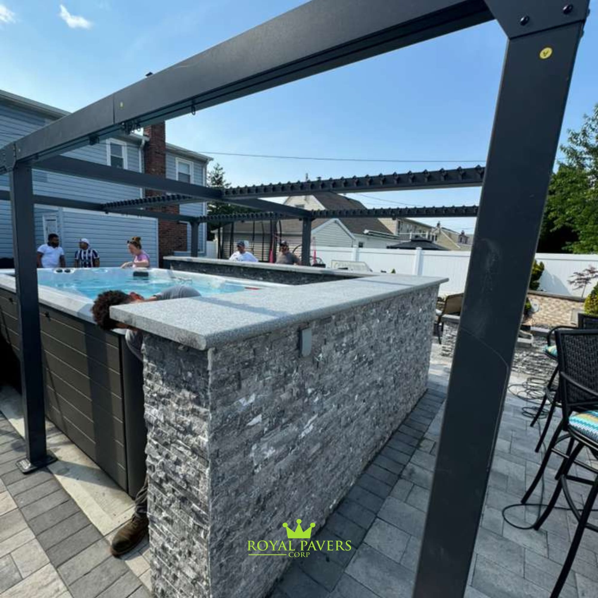Backyard luxury featuring an outdoor kitchen with a stone block base, light paver patio, and swimming pool under a modern pergola, Long Island by Royal Pavers Corp